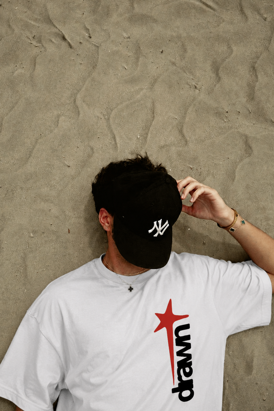 Person wearing a black cap with a white logo and a white t-shirt with a red and black design, against a beige wall.